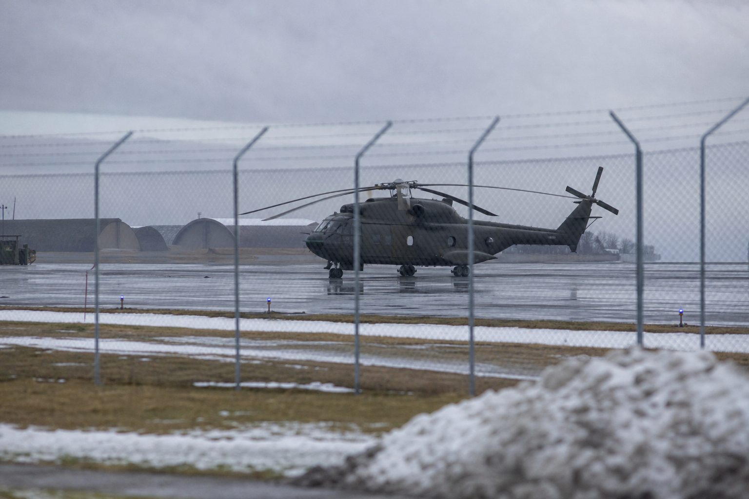 Et militært helikopter står på en norsk flyplass i overskyet vær, sett på avstand gjennom et gjerde.