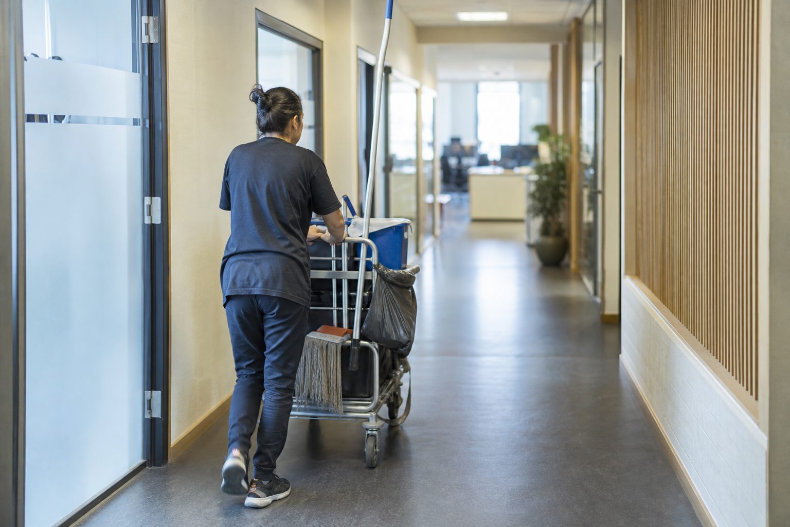 Redaksjonelt foto av en renholder med rengjøringsvogn i korridoren på en kommunal kontorbygning, med arbeidsutstyr og lokalene tydelig synlige.