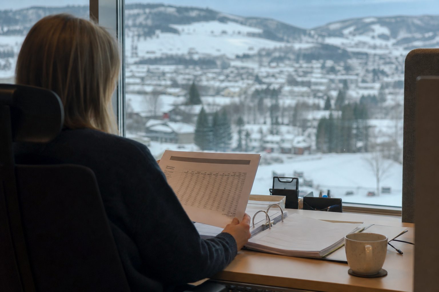 En saksbehandler sitter ved et skrivebord på et norsk fylkeshus og leser et utskrevet brev med en tabell.