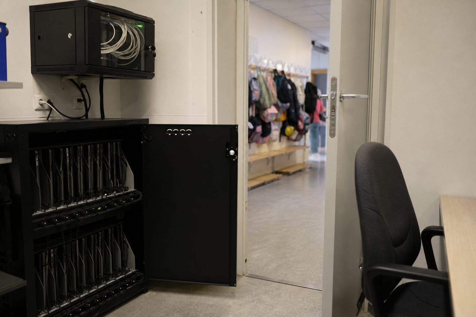 Et lite IKT-rom på en norsk barneskole med stablede bærbare PC-er i ladeskap, en nettverkssvitsj på veggen og en tom kontorstol.