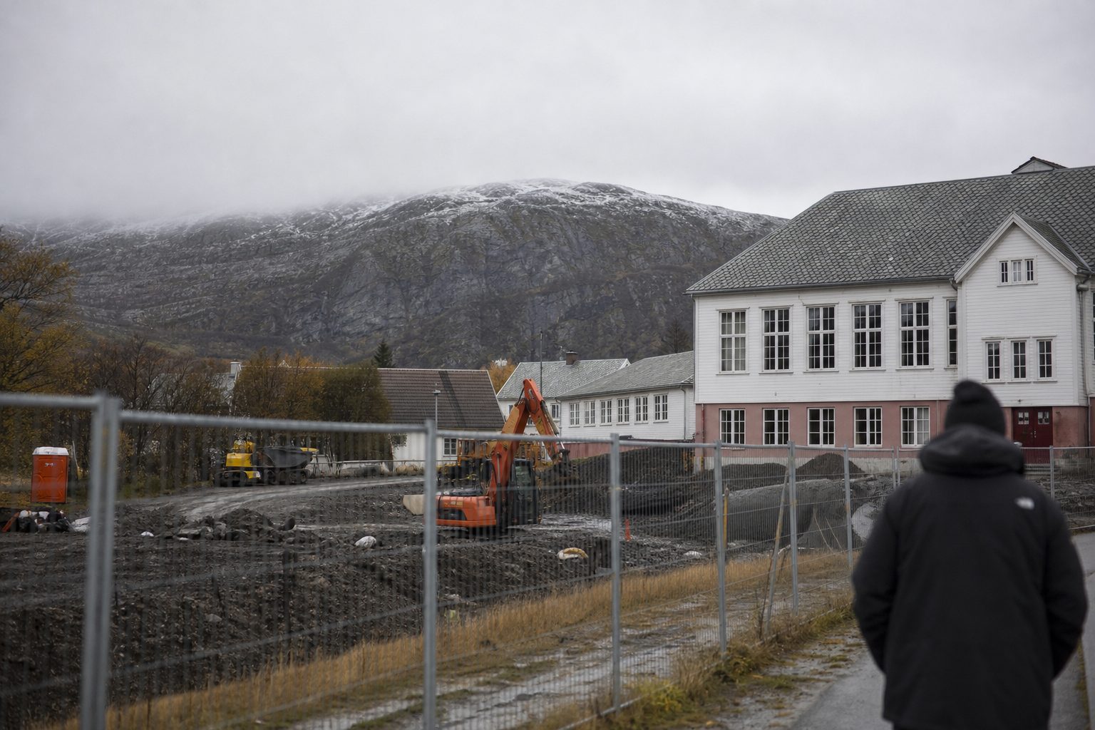 En byggetomt i en liten by i Finnmark en grå høstdag, med inngjerdet område, anleggsmaskiner og byggegjerder ved siden av en eldre skolebygning.