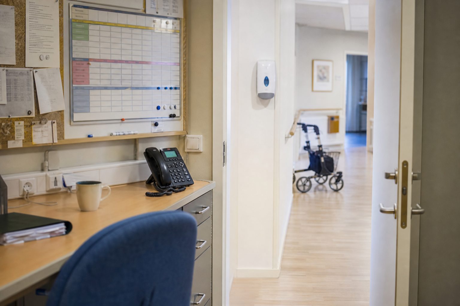 Et vaktrom på et norsk sykehjem med en vaktplan med åpne felt på oppslagstavlen, en telefon og en kaffekopp på pulten.