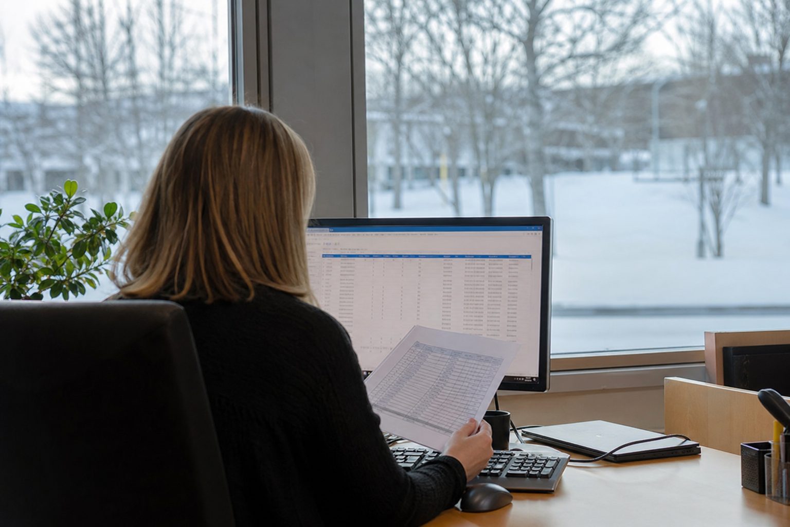 En innkjøper ved et skrivebord i et norsk kommunehus sammenligner en utskrevet brukerliste med bestillinger på en dataskjerm.