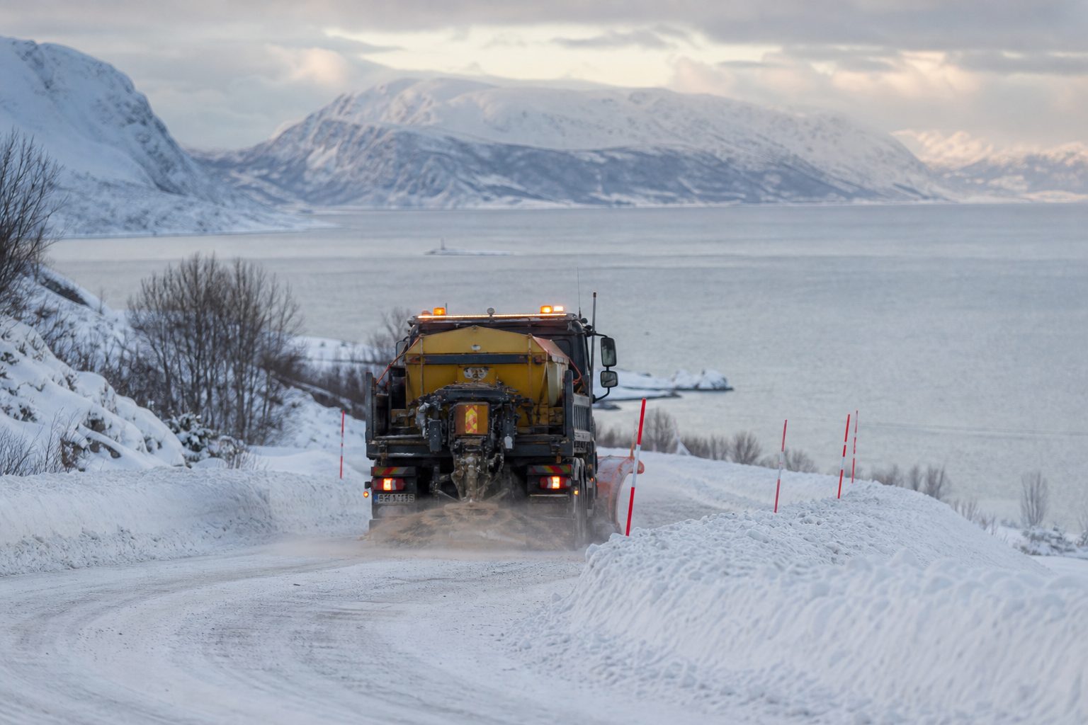En brøytebil strør sand på en snødekt fylkesvei i et kupert nordnorsk kystlandskap.