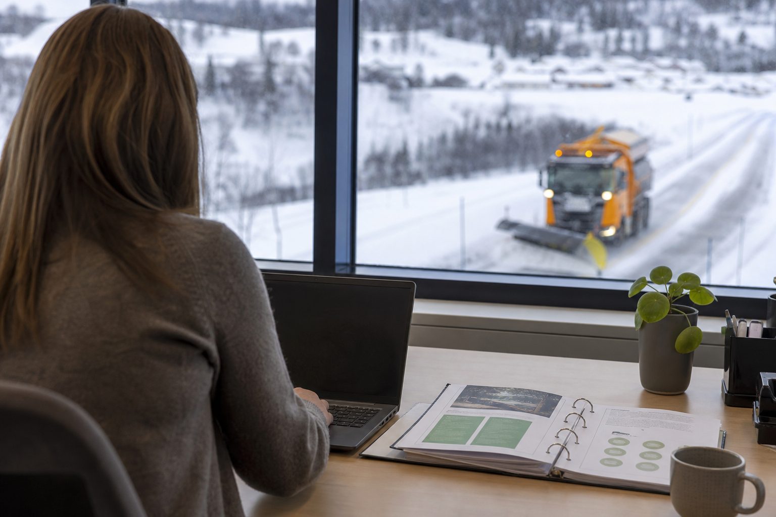 En innkjøper sitter ved et skrivebord på et norsk fylkeskommunalt kontor med en åpen perm med miljørutiner og en bærbar PC.