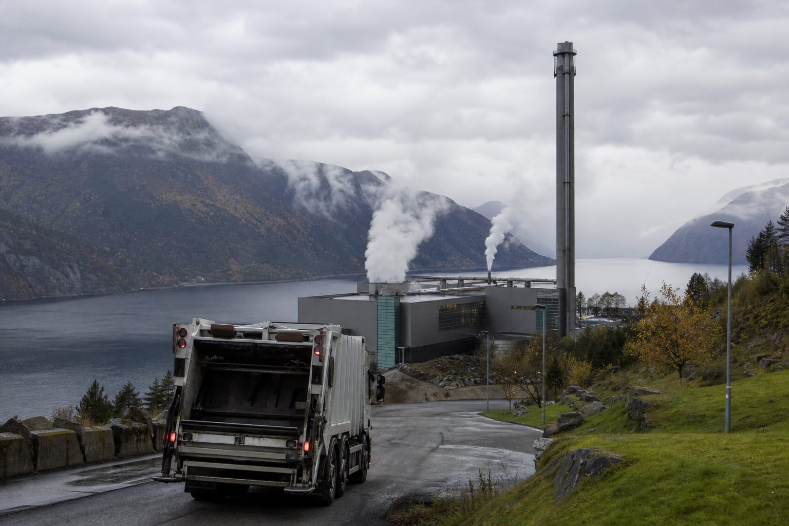 En avfallsbil kjører inn mot et forbrenningsanlegg med høy pipe en grå høstdag på Vestlandet, med fjell og fjord i bakgrunnen.