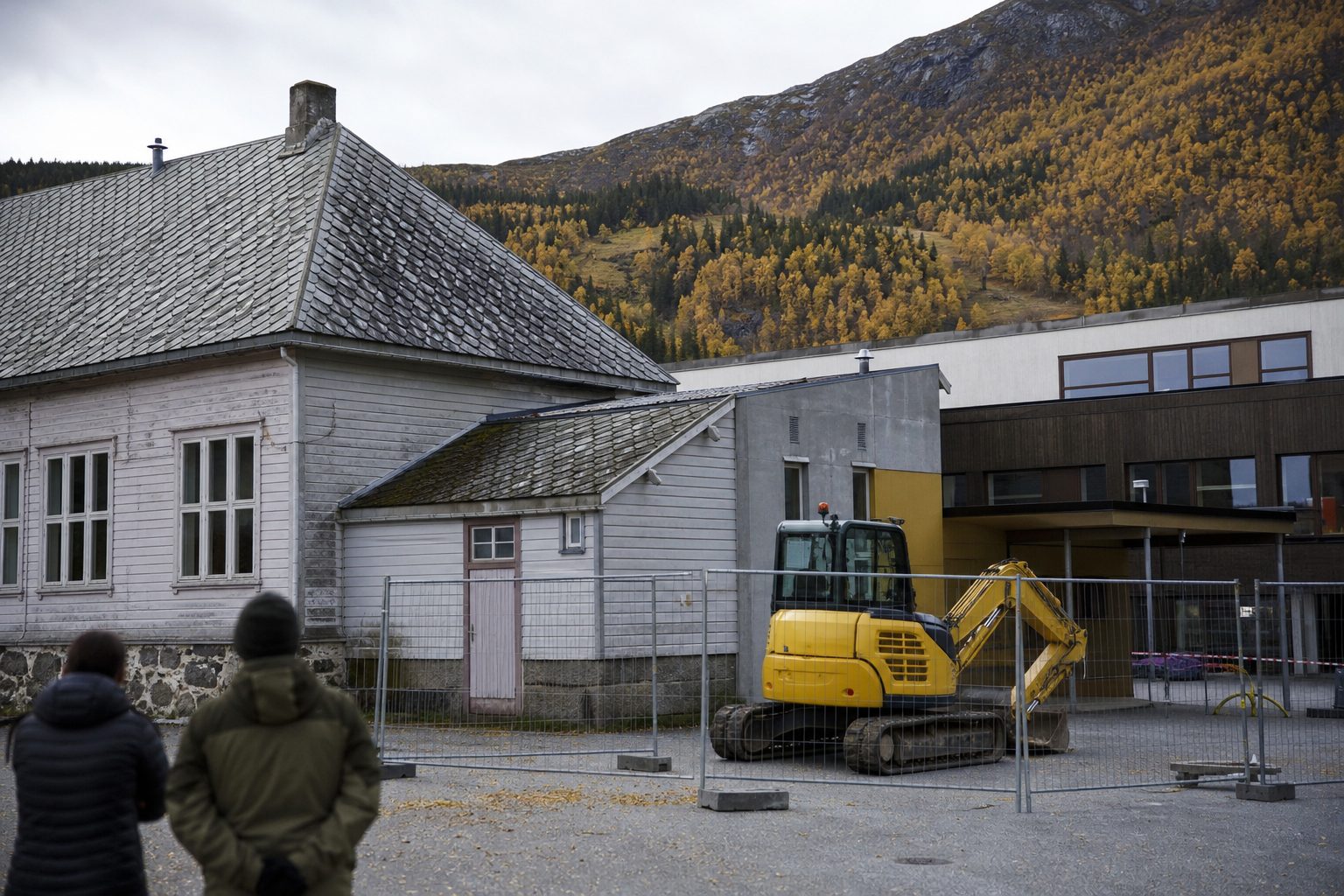 En eldre skolebygning i en norsk bygd om høsten, med en lav, gammel fløy i tre og mur ved siden av en nyere bygningsdel.