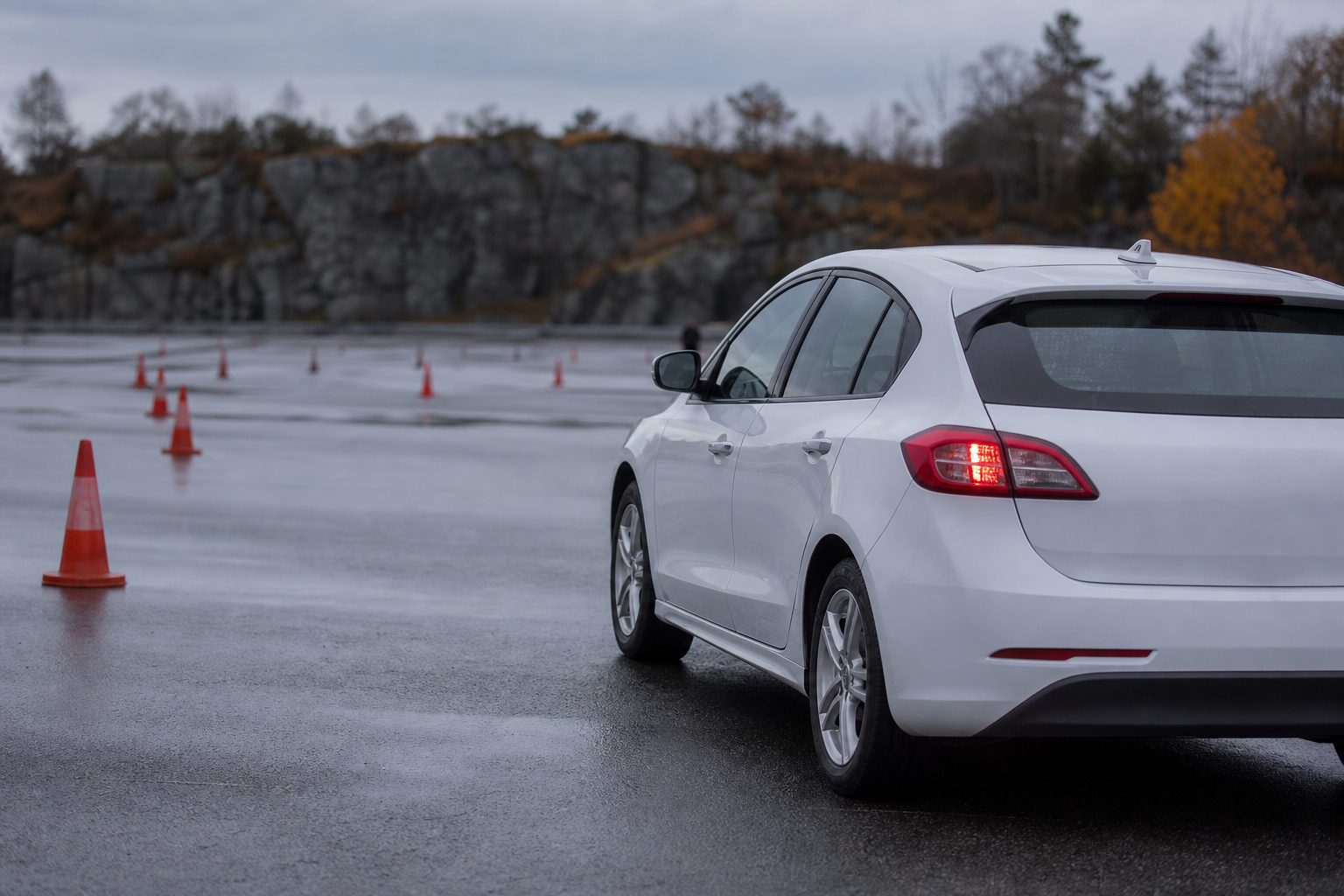 En hvit personbil for kjøreopplæring står parkert på en våt øvingsbane utenfor Stavanger en grå høstdag, med trafikkjegler oppstilt på asfalten foran.